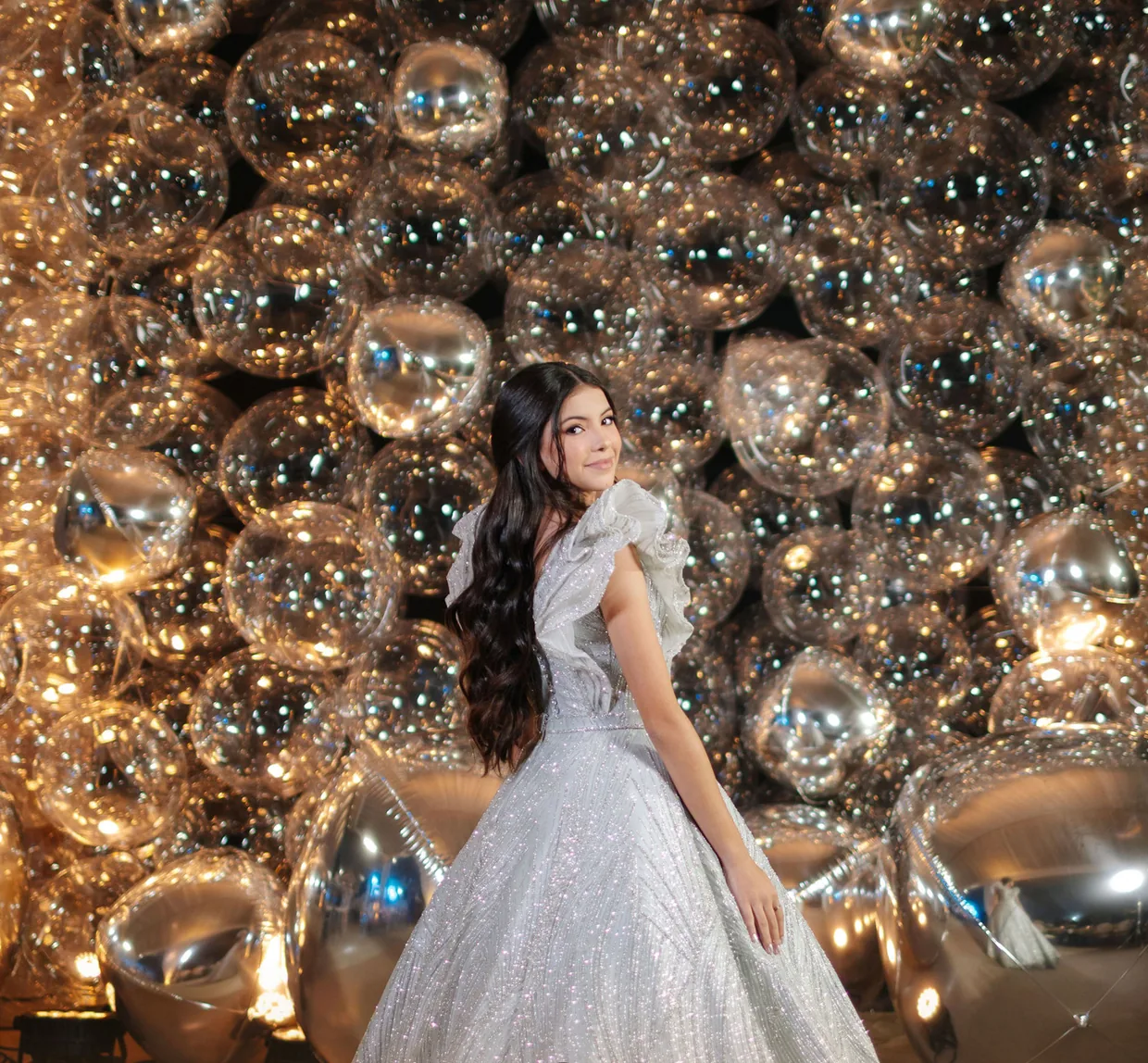 Debutante de vestido prateado diante de uma parede de balões espelhados
